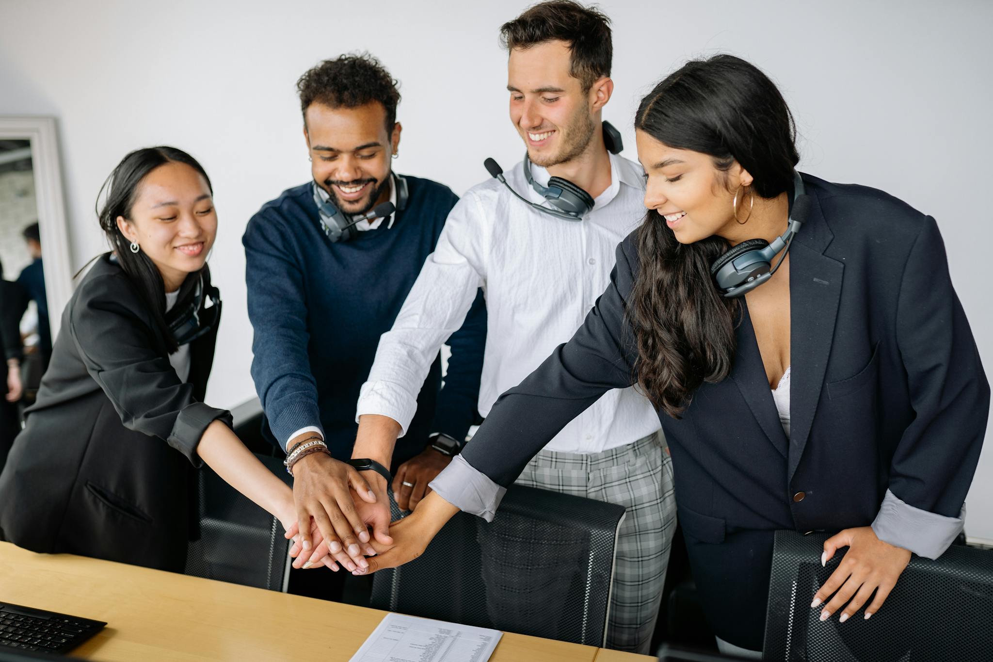 A diverse group of call center agents in an office joining hands in a teamwork gesture, smiling and wearing headsets.